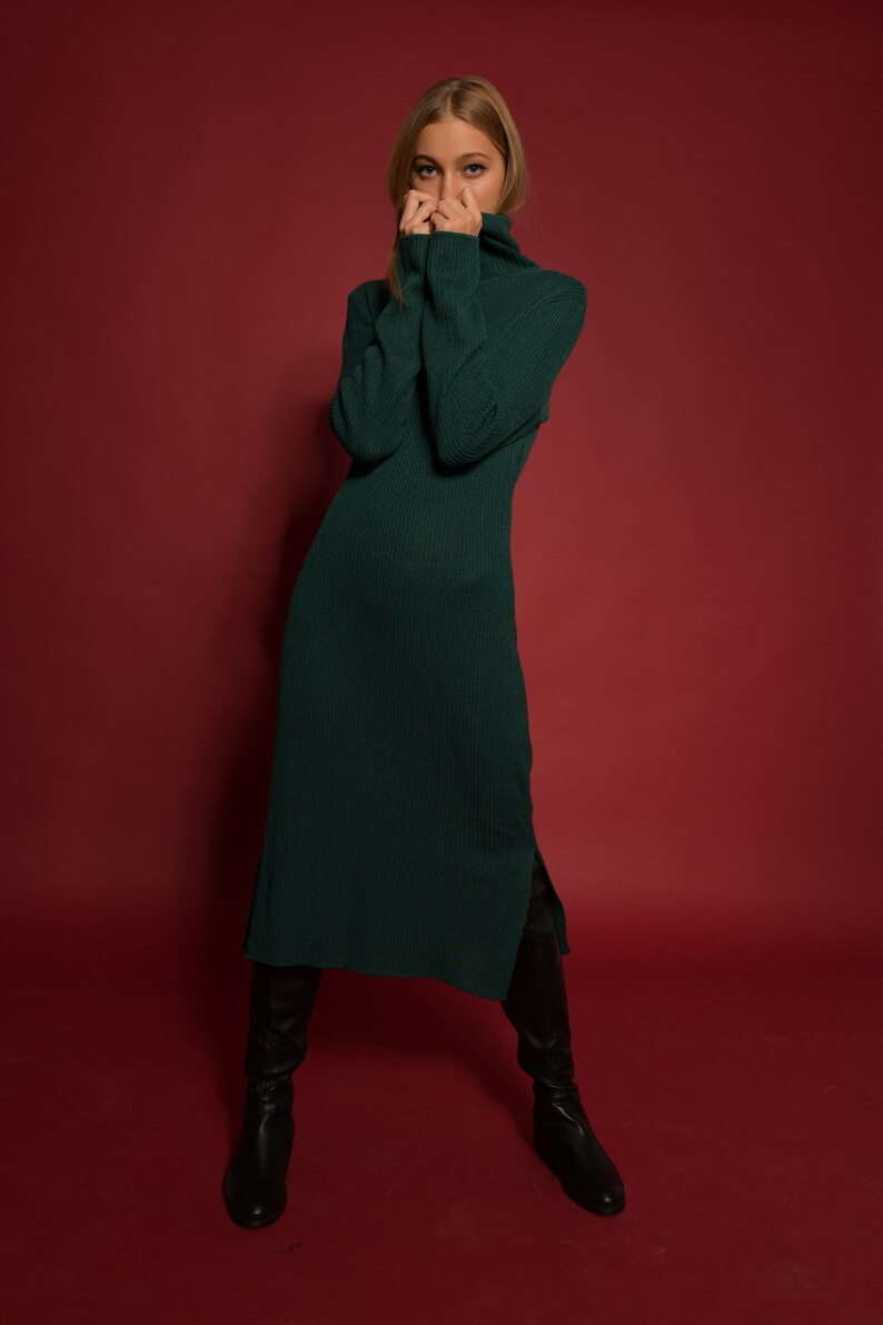 May include: A woman wearing a long, dark green turtleneck sweater dress with a side slit. She is wearing black knee-high boots and standing on a red background.