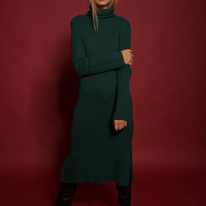 May include: A woman wearing a long, dark green turtleneck sweater dress with a ribbed texture. She is standing on a red background and wearing black knee-high boots.