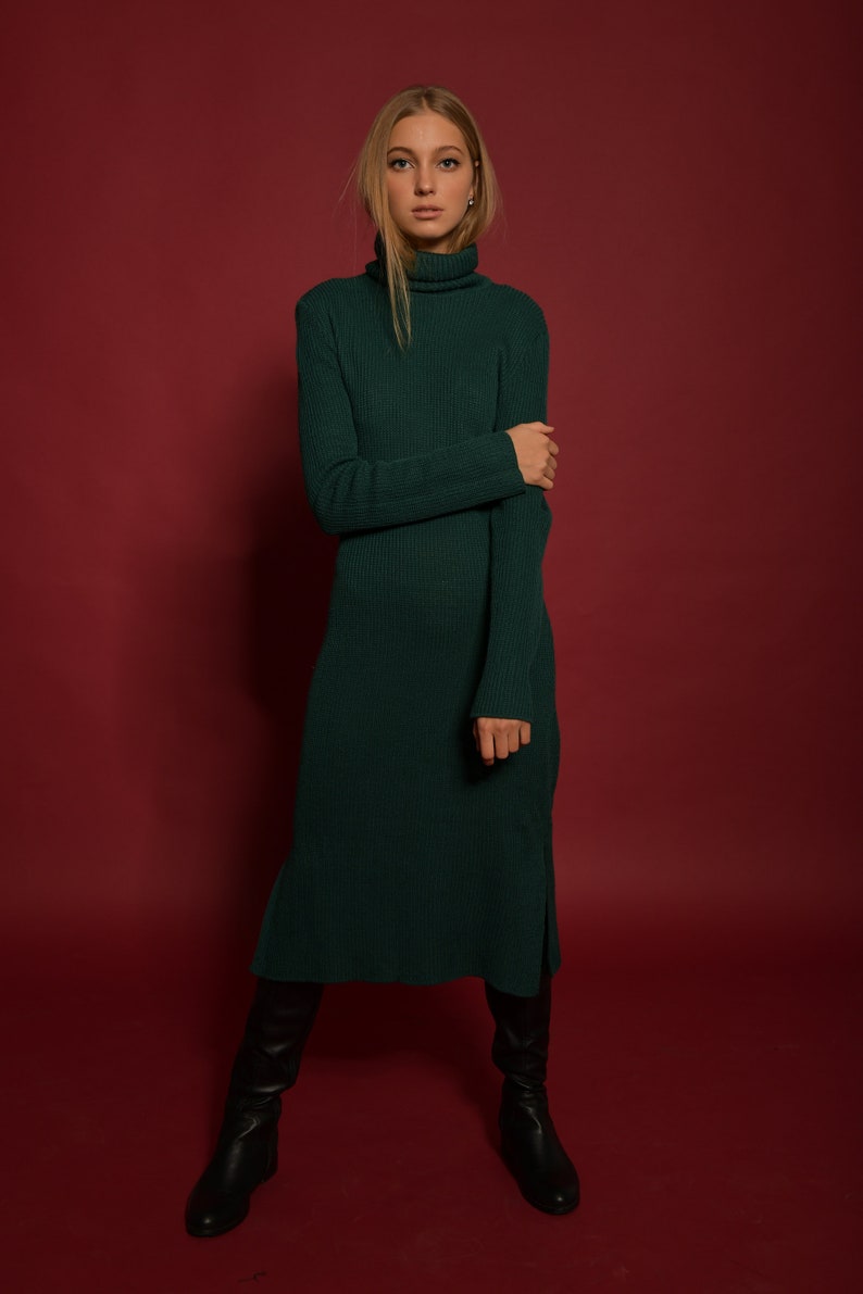 May include: A woman wearing a long, dark green turtleneck sweater dress with a ribbed texture. She is standing on a red background and wearing black knee-high boots.