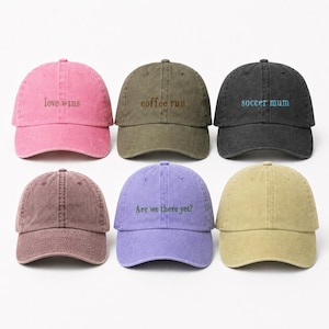 May include: Six baseball caps in various colours including pink, olive green, charcoal, maroon, purple, and tan. Each cap features embroidered text: love wins, coffee run, soccer mum, and Are we there yet? The caps have a washed, vintage appearance.