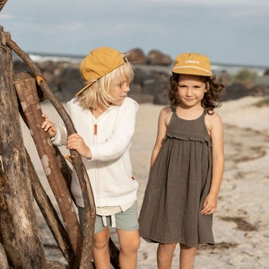 May include: Two children wearing yellow baseball caps stand on a driftwood log. The girl is wearing a dark brown dress and the boy is wearing a white sweater and green shorts. The girl's cap has the text "CLIMB & CO." on it.