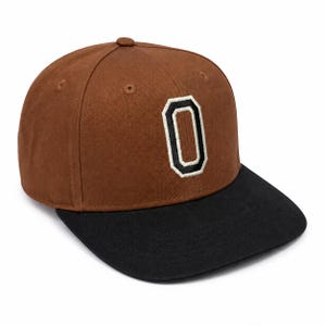 May include: A brown baseball cap with a black brim and a central embroidered letter O. The letter O is stitched in white with a thick black outline, creating a bold contrast against the brown crown of the cap. The cap features six panels.