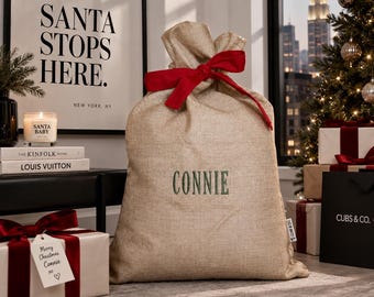 May include: A tan hessian sack with a drawstring top tied with a red ribbon. The name CONNIE is embroidered in dark green thread on the front of the sack. A small white fabric tag with black text is attached to the side seam of the sack.