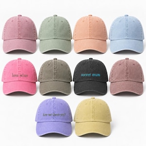 May include: A collection of ten baseball caps in various colours including pink, sage green, peach, light blue, dark grey, and yellow. Some caps feature embroidered text such as love wins, soccer mum, and Are we there yet? in contrasting thread.