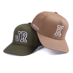 May include: Two baseball caps, one green with "JR" embroidered in white and black, and one brown with "K" embroidered in white and gold. The brown cap has "CUBS & CO." embroidered on the side.