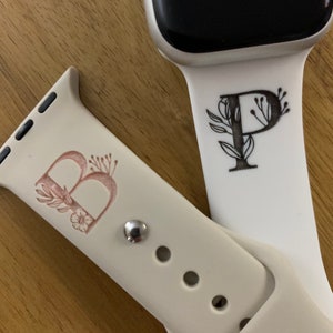 Pode incluir: Duas pulseiras brancas para Apple Watch com gravações personalizadas. Uma pulseira apresenta a letra "B" com detalhes florais em rosa claro. A outra pulseira apresenta a letra "P" com detalhes florais em preto.