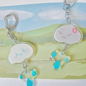 May include: Two white cloud-shaped keychains with blue raindrop charms. One cloud has a frowning face and the other has a happy face with the Japanese characters for "smile".