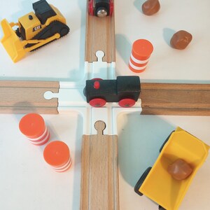 May include: A wooden train track set with a crossing, featuring a yellow bulldozer, two black toy trains with red wheels, orange and white traffic cones, and a yellow dump truck with a brown rock.