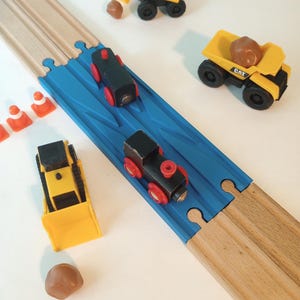 May include: A wooden toy train track with a blue bridge section. Toy trains and construction vehicles, including a yellow excavator and dump truck, are arranged on and around the track. Small orange traffic cones and brown rocks are also present.