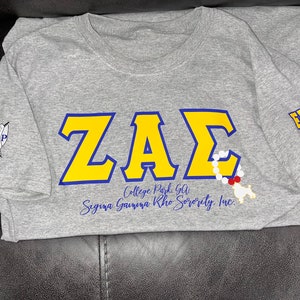 May include: Gray t-shirt with a yellow and blue "ZAE" design, a white and red beaded necklace, and the text "College Park, GA Sigma Gamma Rho Sorority, Inc."  The right sleeve has the text "FALL 2022" in yellow and blue.