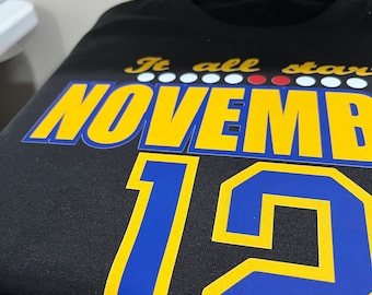 Pode incluir: Camiseta preta com texto amarelo e azul que diz "It all starts November 12th".