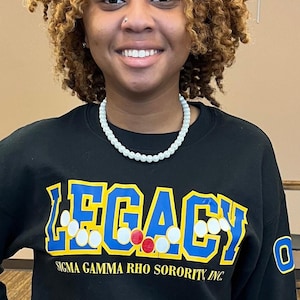 May include: A black long-sleeve sweatshirt featuring the word LEGACY in large blue letters with yellow outlines. The text includes white and red circular accents. Below, the text reads SIGMA GAMMA RHO SORORITY, INC. in yellow font.