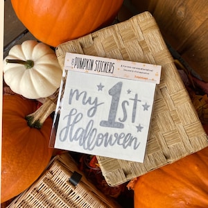 May include: A sheet of vinyl pumpkin stickers with the text "my 1st Halloween" in grey script, surrounded by stars. The stickers are displayed on a woven basket, with pumpkins in the background.
