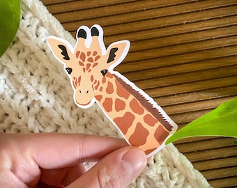 May include: A brown and tan giraffe sticker with a white background. The giraffe's head is facing the left side of the image. The sticker is being held by a hand.