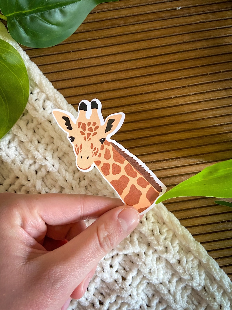 May include: A brown and tan giraffe sticker with a white background. The giraffe's head is facing the left side of the image. The sticker is being held by a hand.