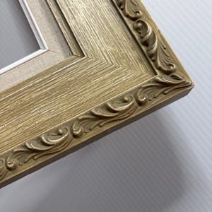 May include: Close-up of a gold-colored picture frame with ornate, carved detailing. The frame has a textured surface and a neutral-colored canvas backing. The corner of the frame is visible, showcasing the intricate design.