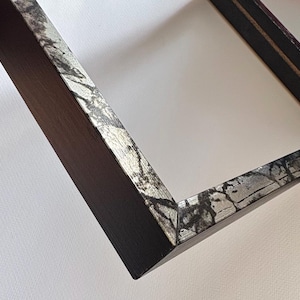 May include: A dark brown picture frame with a silver and black marbled design on the outer edge. The frame is angled, showing the corner and the inside edge against a white background.