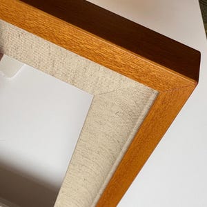 May include: A rectangular picture frame featuring a warm-toned, polished wooden outer border with a mitered corner joint. The inner section of the frame is lined with a textured, light beige fabric border that creates a neutral matting effect.
