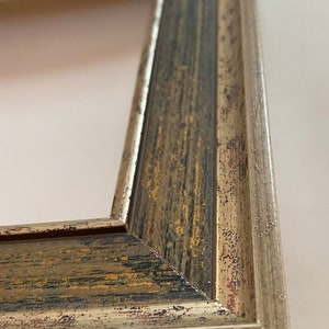 May include: A close-up of a picture frame with a distressed, aged, silver and gold finish. The frame has a raised edge and a decorative pattern.