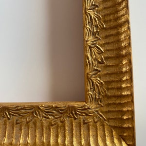 May include: Close-up of a gold-colored picture frame. The frame has a textured, ridged design with a decorative leaf pattern along the inner edge. The frame's corner is visible, showing the mitered joint. The frame is set against a white background.