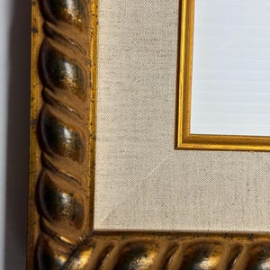 May include: Close-up of a decorative gold picture frame with a textured, rope-like design around the edges. The frame surrounds a canvas-like material and a white space, suggesting a space for a picture or artwork.