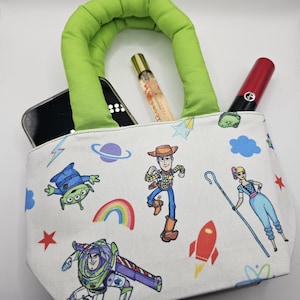 May include: A small, white tote bag with a green handle, featuring colorful cartoon character graphics. The bag contains a smartphone, perfume, and a red lipstick. The bag's design includes images of a rainbow, stars, and a rocket.