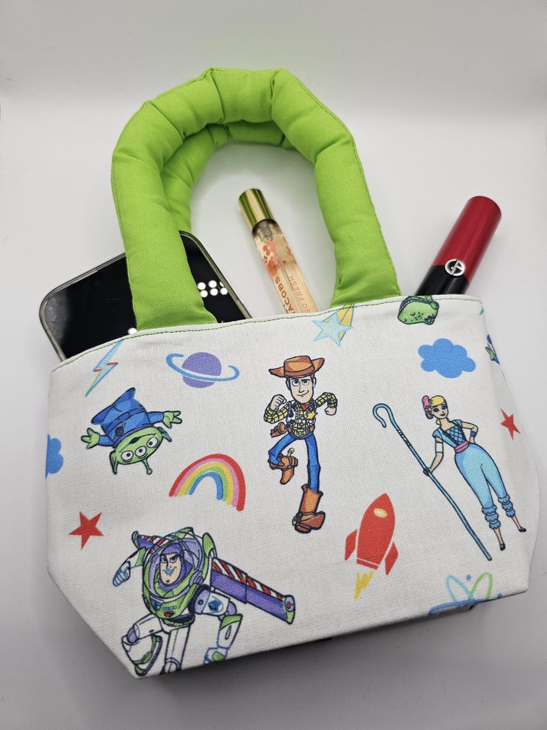 May include: A small, white tote bag with a green handle, featuring colorful cartoon character graphics. The bag contains a smartphone, perfume, and a red lipstick. The bag's design includes images of a rainbow, stars, and a rocket.