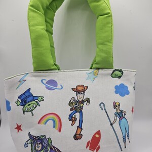 May include: A small, white tote bag with a green handle. The bag features colourful cartoon characters, including a cowboy, a space ranger, and an alien. A rainbow, stars, and a rocket are also printed on the bag.