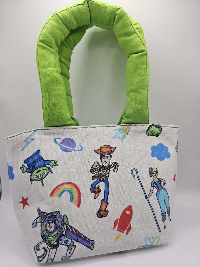 May include: A small, white tote bag with a green handle. The bag features colourful cartoon characters, including a cowboy, a space ranger, and an alien. A rainbow, stars, and a rocket are also printed on the bag.