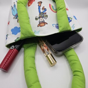 May include: A green-handled tote bag with a white lining featuring a colorful cartoon print. The bag contains a smartphone, a red lipstick, and a gold-capped perfume bottle. The bag's handles are a bright green.