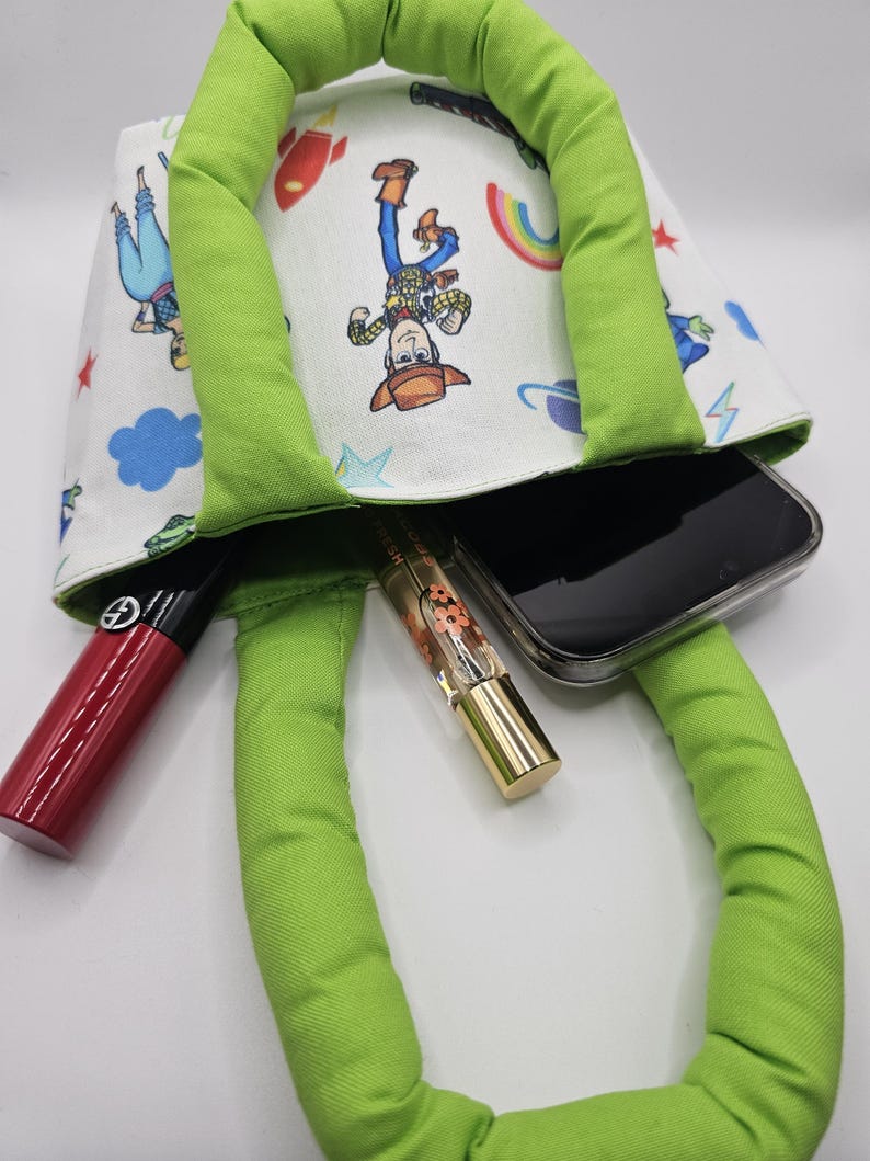 May include: A green-handled tote bag with a white lining featuring a colorful cartoon print. The bag contains a smartphone, a red lipstick, and a gold-capped perfume bottle. The bag's handles are a bright green.