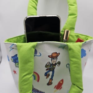 May include: A small, white tote bag with a green handle and interior lining. The bag features a colourful print of cartoon characters, including a cowboy. A smartphone and lipstick are visible inside the bag.