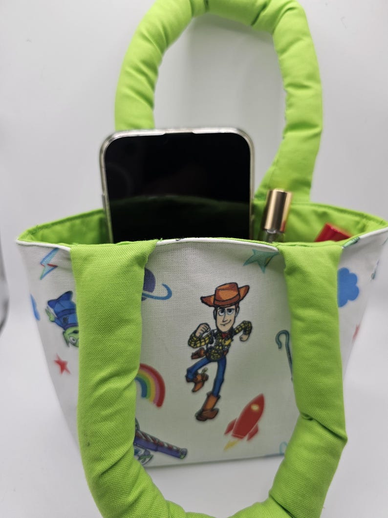 May include: A small, white tote bag with a green handle and interior lining. The bag features a colourful print of cartoon characters, including a cowboy. A smartphone and lipstick are visible inside the bag.
