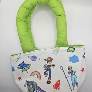 May include: A small white tote bag with a green padded handle. The bag is decorated with colourful cartoon characters, including Woody, Buzz Lightyear, and an alien, along with a rainbow, stars, and a rocket ship.