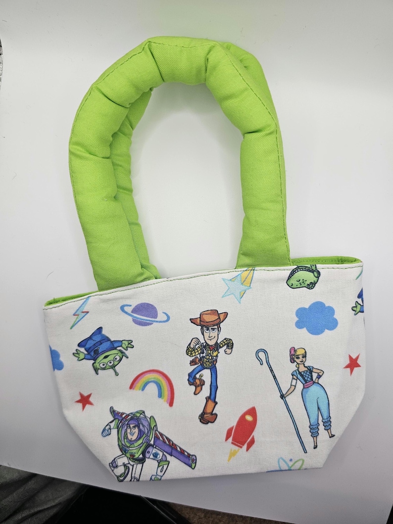 May include: A small white tote bag with a green padded handle. The bag is decorated with colourful cartoon characters, including Woody, Buzz Lightyear, and an alien, along with a rainbow, stars, and a rocket ship.