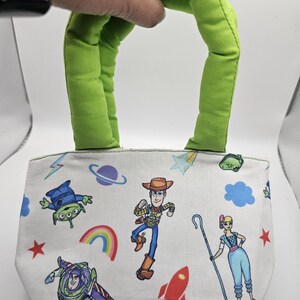 May include: A small, white tote bag with a green handle, decorated with cartoon characters. The bag features a repeating pattern of characters, a rainbow, a rocket, and stars. The handle is a bright green.