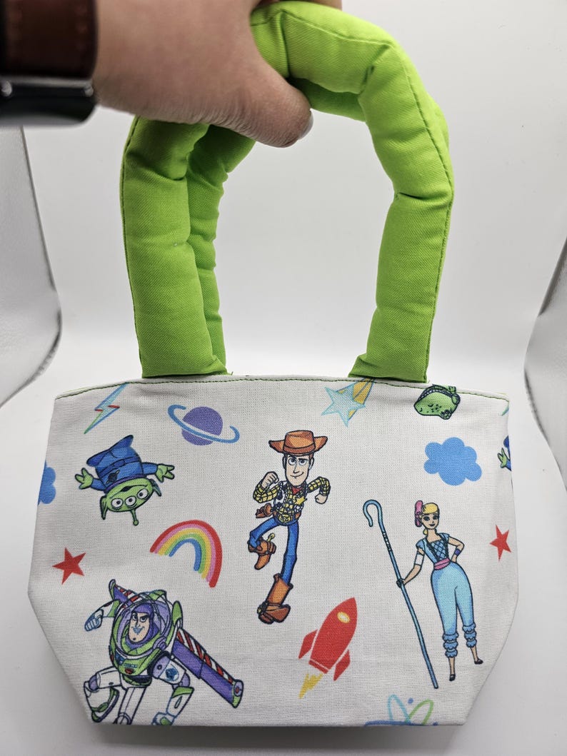May include: A small, white tote bag with a green handle, decorated with cartoon characters. The bag features a repeating pattern of characters, a rainbow, a rocket, and stars. The handle is a bright green.