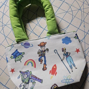 May include: A small, white tote bag featuring colorful cartoon characters and space-themed graphics. The bag has a green padded handle and is decorated with images of Woody, Buzz Lightyear, and other characters. The bag is approximately 20 cm wide.