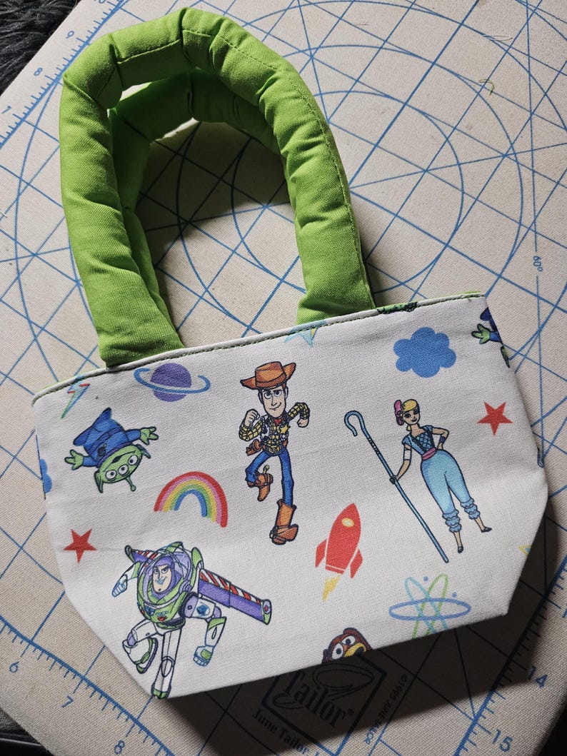 May include: A small, white tote bag featuring colorful cartoon characters and space-themed graphics. The bag has a green padded handle and is decorated with images of Woody, Buzz Lightyear, and other characters. The bag is approximately 20 cm wide.