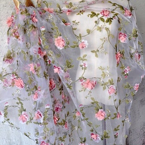 Puede incluir: Una tela blanca transparente con un patrón floral con rosas rosas y hojas verdes. La tela tiene una calidad delicada, casi translúcida.