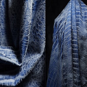 May include: Close-up of a draped fabric with a crocodile skin pattern. The fabric is a deep blue colour with a lighter, textured pattern overlaying it. The folds and shadows create depth and dimension.