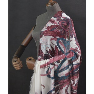 Pode incluir: Um tecido branco com um padrão paisley em tons de bordô, verde-azulado e azul claro é drapeado sobre um manequim. O tecido tem uma aparência delicada e fluida, com uma borda com franjas visível na parte inferior. O design paisley apresenta elementos florais e abstratos intrincados.