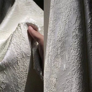 May include: Close-up of a white fabric with a textured, bumpy surface. The fabric has a subtle sheen and appears to be soft and luxurious.