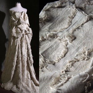 Puede incluir: Un vestido blanco hecho de una tela texturizada con bordes deshilachados. La tela está recogida y drapeada para crear una silueta fluida.