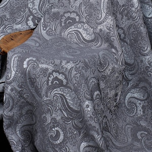 K&ouml;nnte beinhalten: Nahaufnahme eines grauen Stoffes mit detailliertem Paisley-Muster. Der Stoff hat einen subtilen Glanz, wobei hellere graue Highlights die wirbelnden Blumen- und Tropfenmotive hervorheben. Der Stoff ist &uuml;ber einen Korbstuhl drapiert.