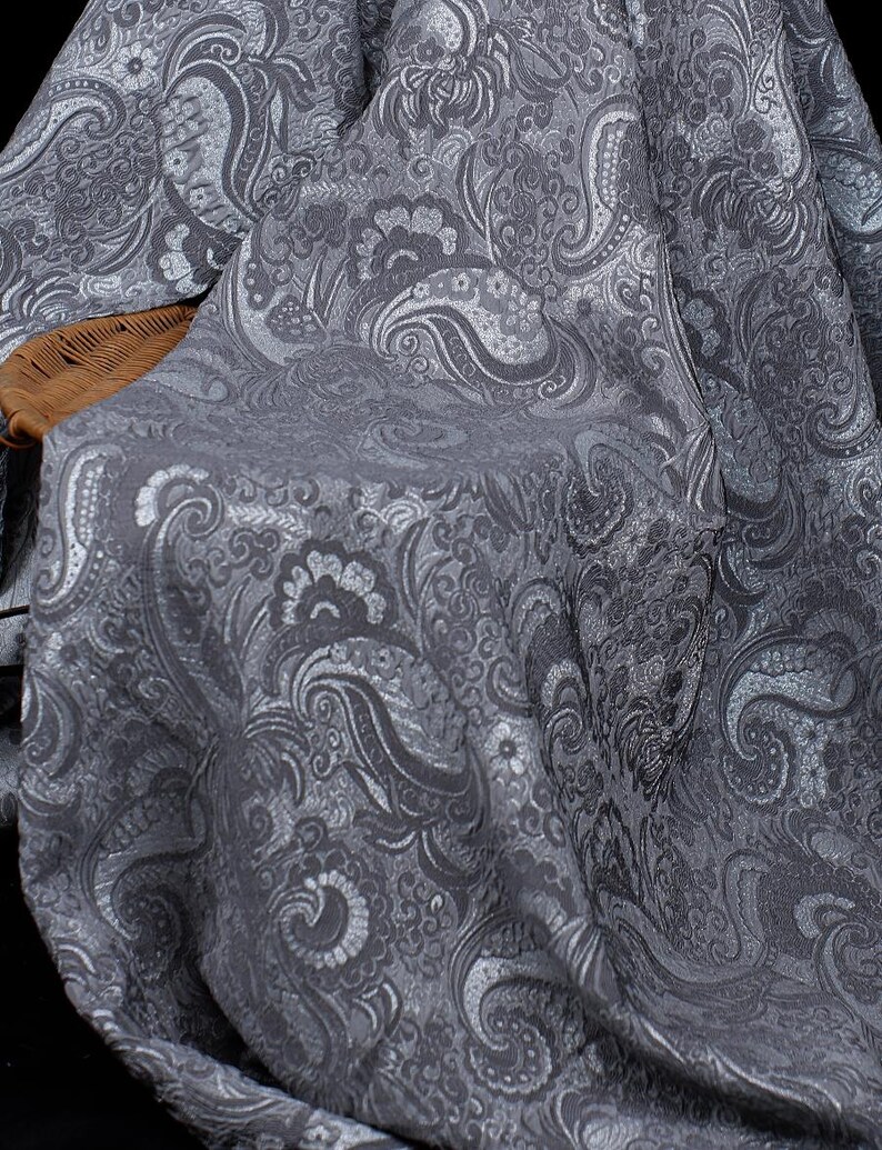 K&ouml;nnte beinhalten: Nahaufnahme eines grauen Stoffes mit detailliertem Paisley-Muster. Der Stoff hat einen subtilen Glanz, wobei hellere graue Highlights die wirbelnden Blumen- und Tropfenmotive hervorheben. Der Stoff ist &uuml;ber einen Korbstuhl drapiert.