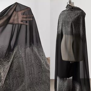 May include: A black and grey cape with a gradient design. The fabric transitions from solid black to speckled grey. The cape is draped over a mannequin, showcasing its flowing silhouette. The fabric is likely a lightweight material.