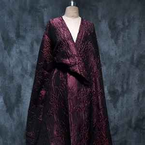 May include: A maroon, long-sleeved, wrap-style dress with a textured, embossed pattern. The dress is displayed on a mannequin against a grey background.