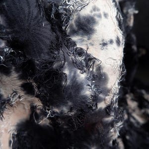 May include: Close-up of a textile with a tie-dye pattern in shades of black, gray, and white. The fabric has a distressed look with frayed edges and loose threads, creating a textured appearance.