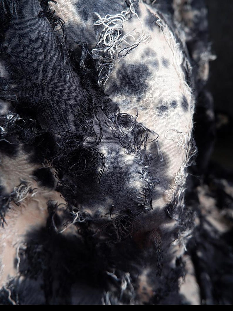 May include: Close-up of a textile with a tie-dye pattern in shades of black, gray, and white. The fabric has a distressed look with frayed edges and loose threads, creating a textured appearance.